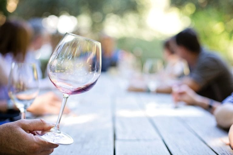 A close-up of a hand holding a wine glass with red wine residue, with people sitting around an outdoor wooden table in the background.