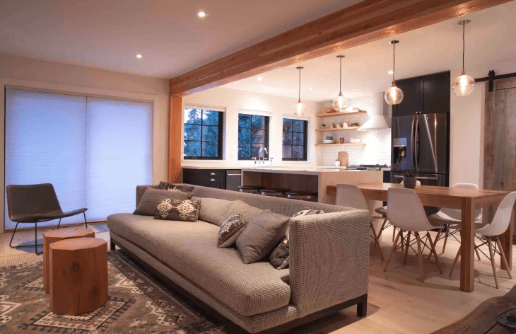 Modern open-plan living room and kitchen with a gray sofa, patterned rug, dining table with white chairs, wooden beams, and pendant lights. Large windows and soft lighting are present.