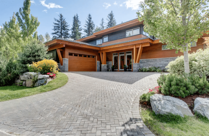 Modern two-story house with wood and stone exterior, large windows, and a paved driveway, surrounded by landscaped plants and trees—perfect for those seeking luxury Airbnb's in Whistler.