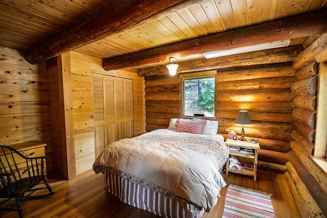 A cozy log cabin bedroom with wooden walls and ceiling, a bed with white bedding, a bedside table with a lamp, a window, a chair, and a small rug on a wooden floor.