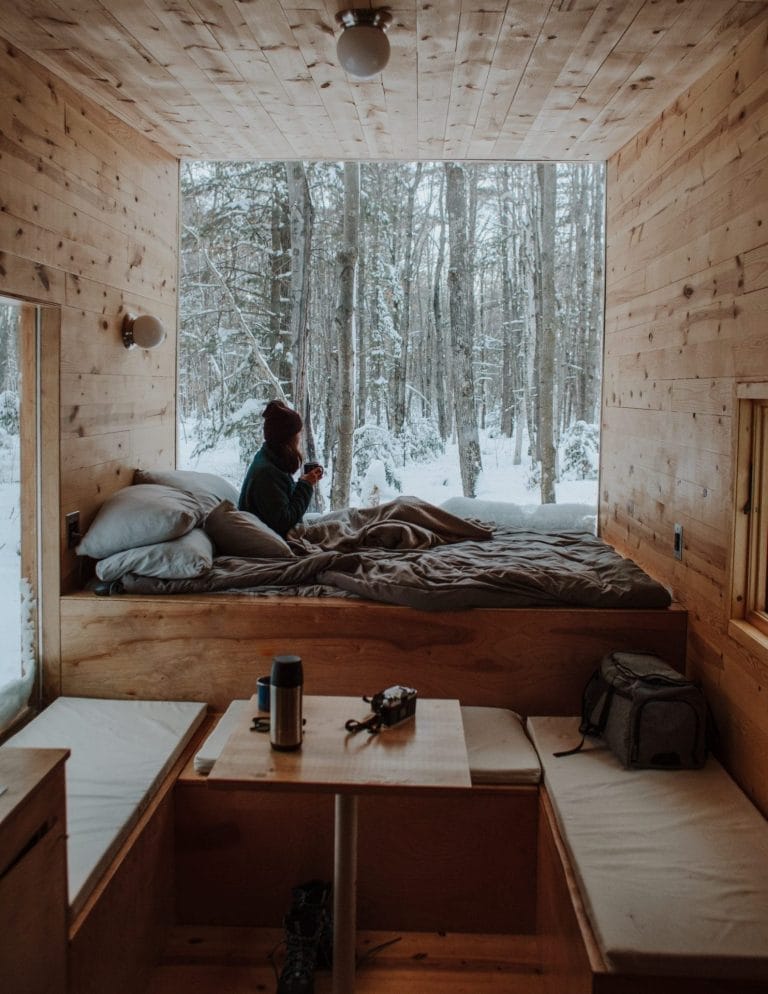 Person sitting on a bed in a wooden cabin, looking out a large window at a snowy forest; a table with mugs, a thermos, and a bag are in the foreground.