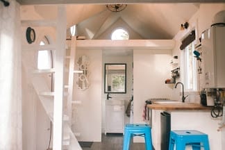 Interior of a small, modern BC Tiny House Airbnb with white walls, loft space, wooden countertops, and two blue stools at a kitchen counter.