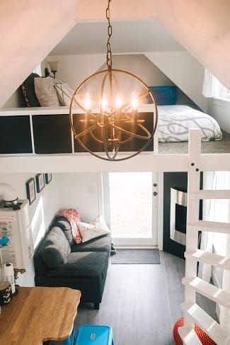 View of a small loft apartment interior, perfect for a BC Tiny House Airbnb, featuring a bed on the upper level, a cozy couch and table below, and a modern hanging light fixture.