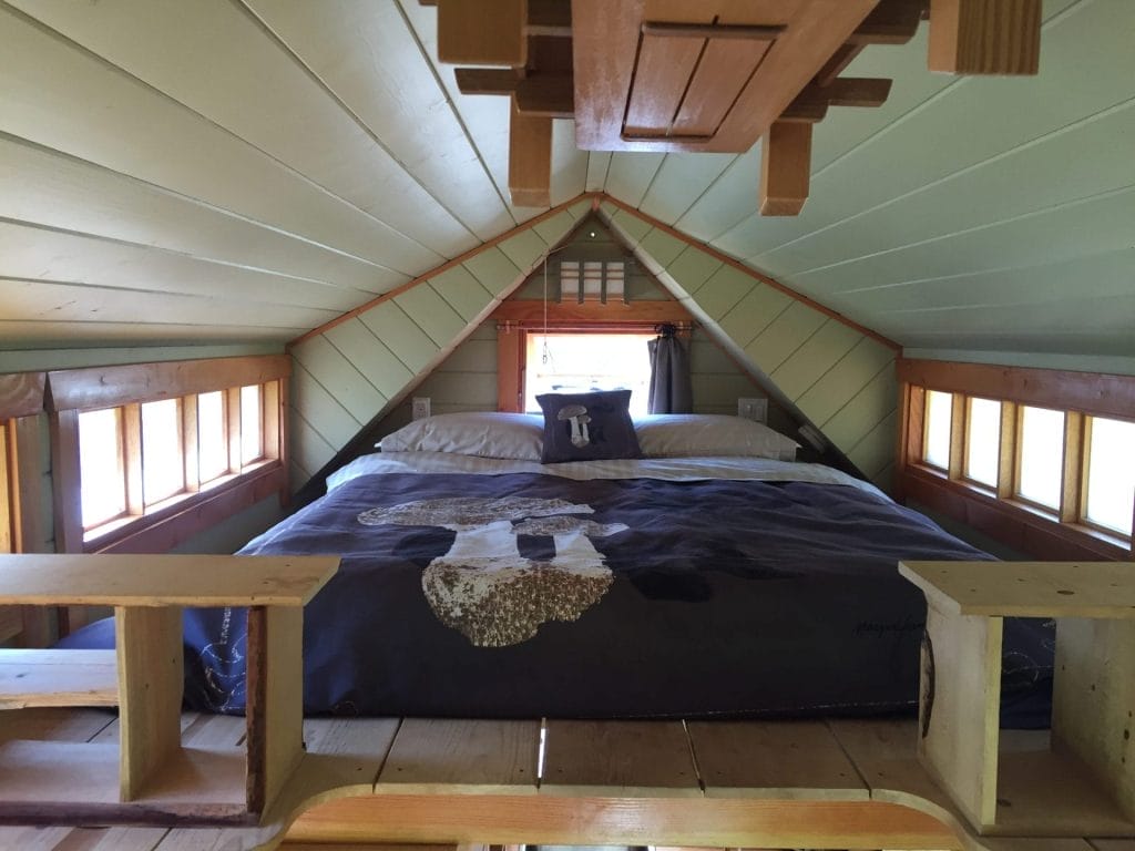 A cozy loft bedroom in this BC Tiny House Airbnb features a low, peaked ceiling, wood paneling, and a double bed; small windows line the side walls, letting in natural light.