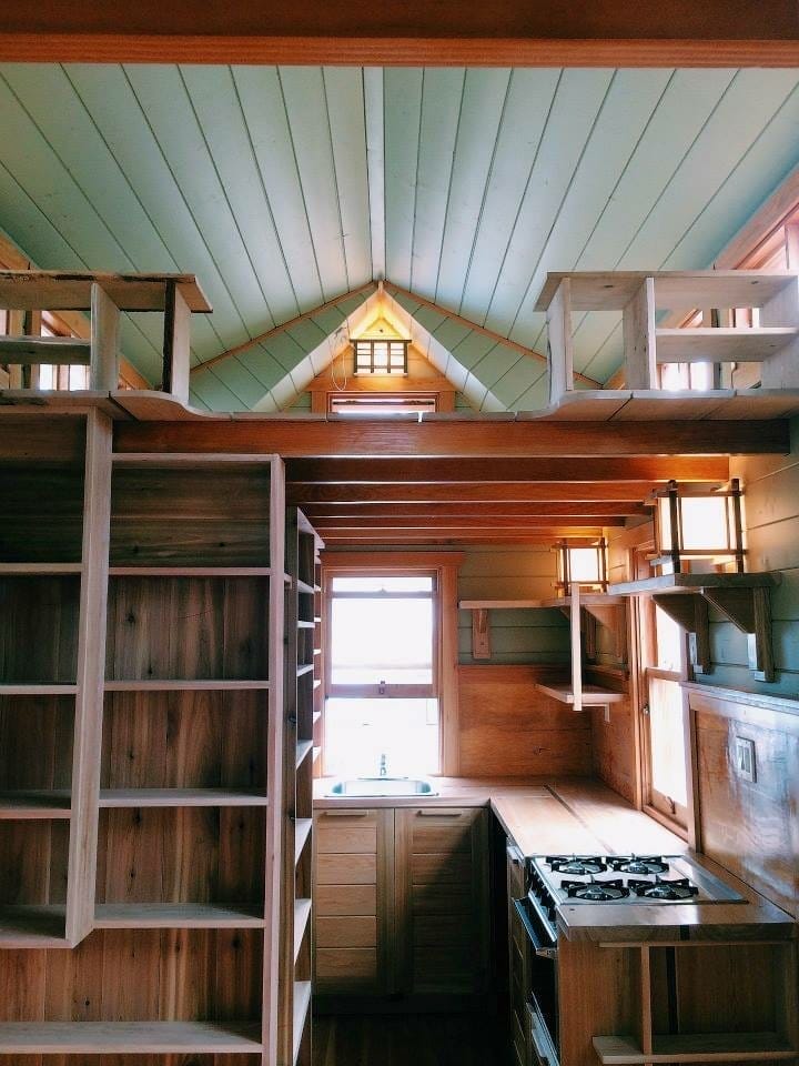 Interior of a small wooden kitchen in a BC Tiny House Airbnb, with a stove and sink below a lofted sleeping area, wooden shelves, and natural light streaming through windows.