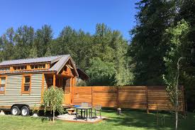 A small wooden BC Tiny House Airbnb on wheels sits on grass near a wooden fence and outdoor table, surrounded by trees under a clear blue sky.