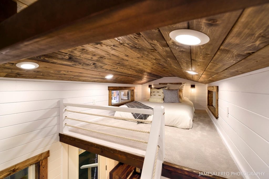 A cozy loft bedroom in this BC Tiny House Airbnb features a low, sloped wooden ceiling, white shiplap walls, a bed with pillows, and a rope railing overlooking the space below.