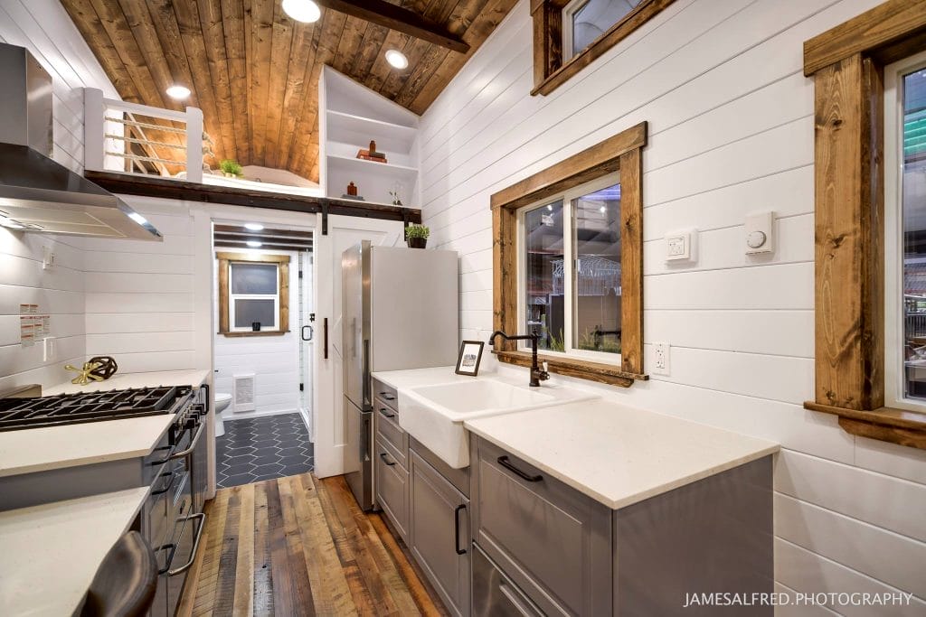 Modern kitchen in a BC Tiny House Airbnb with white shiplap walls, wood accents, farmhouse sink, stainless steel appliances, and a cozy loft area above.