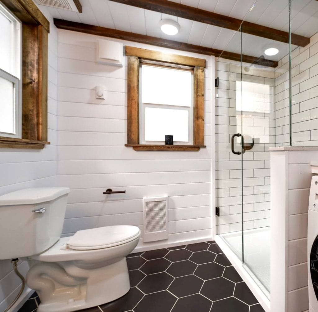 Modern bathroom in this BC Tiny House Airbnb features white shiplap walls, black hexagon floor tiles, a toilet, and a glass-enclosed subway-tiled shower. Two windows with wood trim fill the space with natural light.