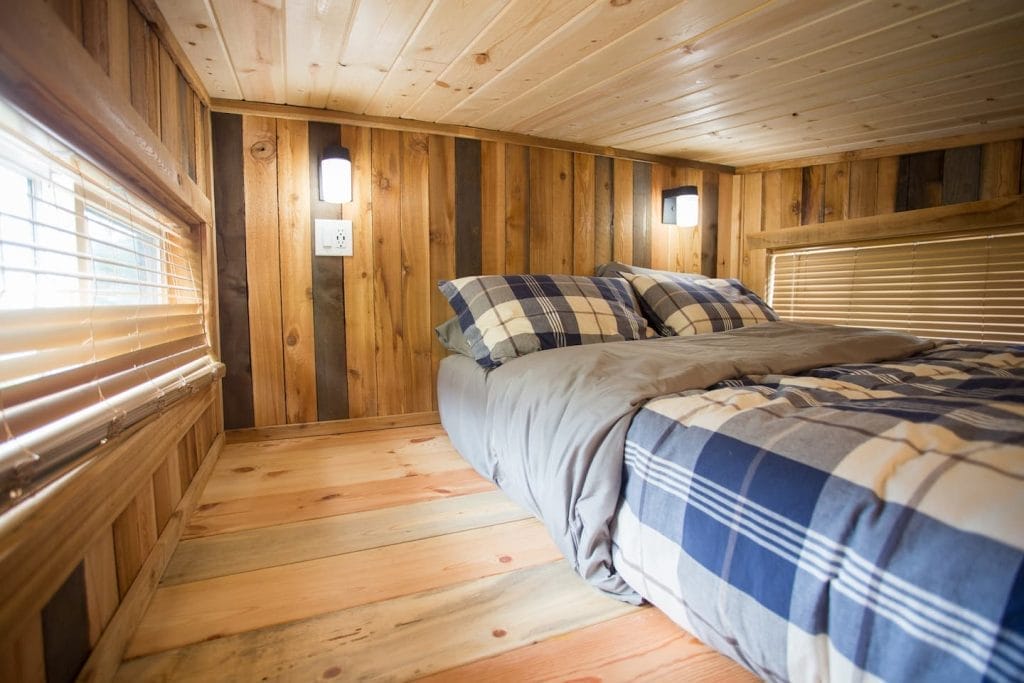 A small, wood-paneled room in a BC Tiny House Airbnb features a low ceiling, plaid-patterned bed, two pillows, and wall-mounted lights beside windows with blinds.