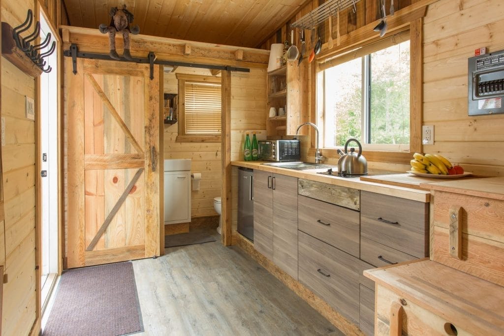 A small, modern kitchen in this BC Tiny House Airbnb features wooden walls, a large window, sleek cabinets, a sink, kettle, fresh fruit on the counter, and a stylish sliding barn door leading to the bathroom.
