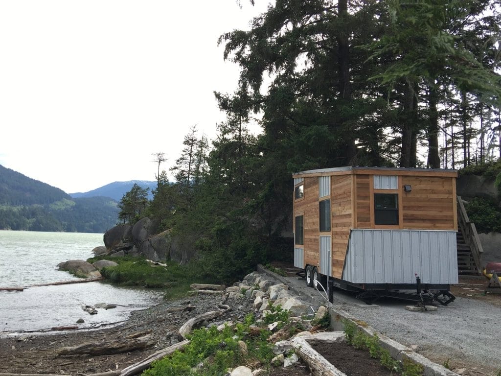 A small wooden BC Tiny House Airbnb on wheels is parked near a rocky shoreline, framed by trees, mountains, and sparkling water in the background.