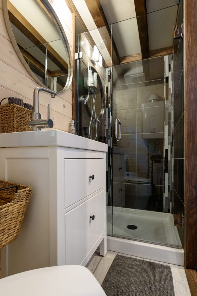 Modern bathroom in a BC Tiny House Airbnb featuring a white vanity and sink, wicker baskets, glass-enclosed shower, and wooden accents on the walls and ceiling.