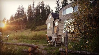 A small wooden BC Tiny House Airbnb with large windows sits in a grassy clearing, surrounded by trees at sunset. Outdoor chairs and a table are arranged near the entrance for relaxing in nature.
