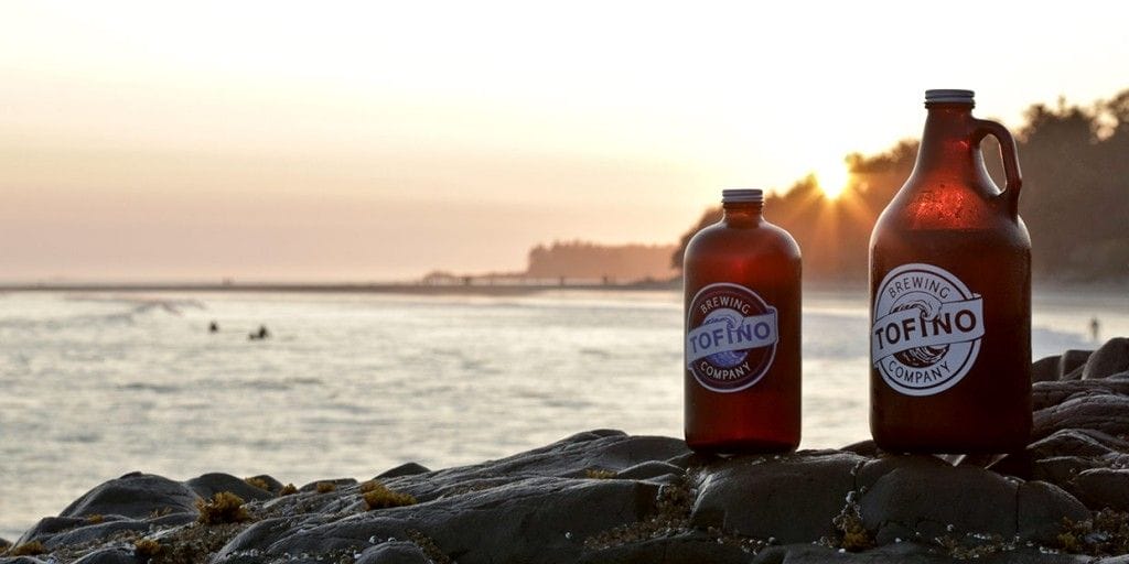 Two brown glass growlers labeled "Tofino Brewing Company" sit on rocks by the ocean at sunset, with surfers in the water and trees in the background, capturing the coastal spirit of Tofino and Ucluelet Breweries.