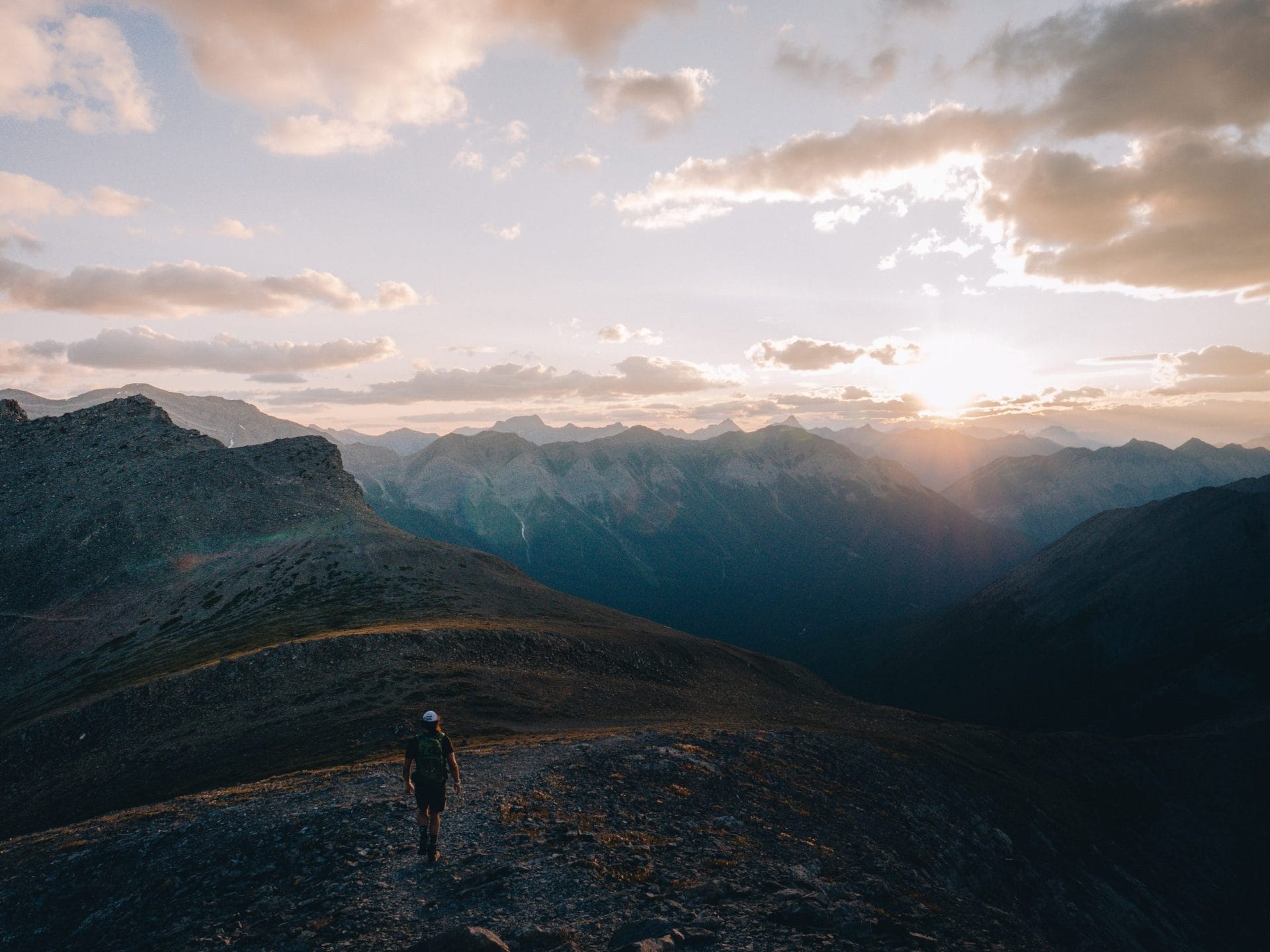 A person with a backpack walks on a mountain ridge at sunset, layers of mountains and scattered clouds in the background—one of the unforgettable things to do in Canmore.
