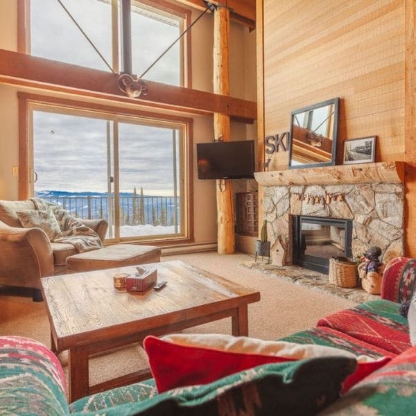 A cozy living room with a stone fireplace, wood beams, and large windows overlooking a balcony with a scenic outdoor view—perfect for relaxing with friends while planning your next Sea to Sky Brewery Guide adventure.