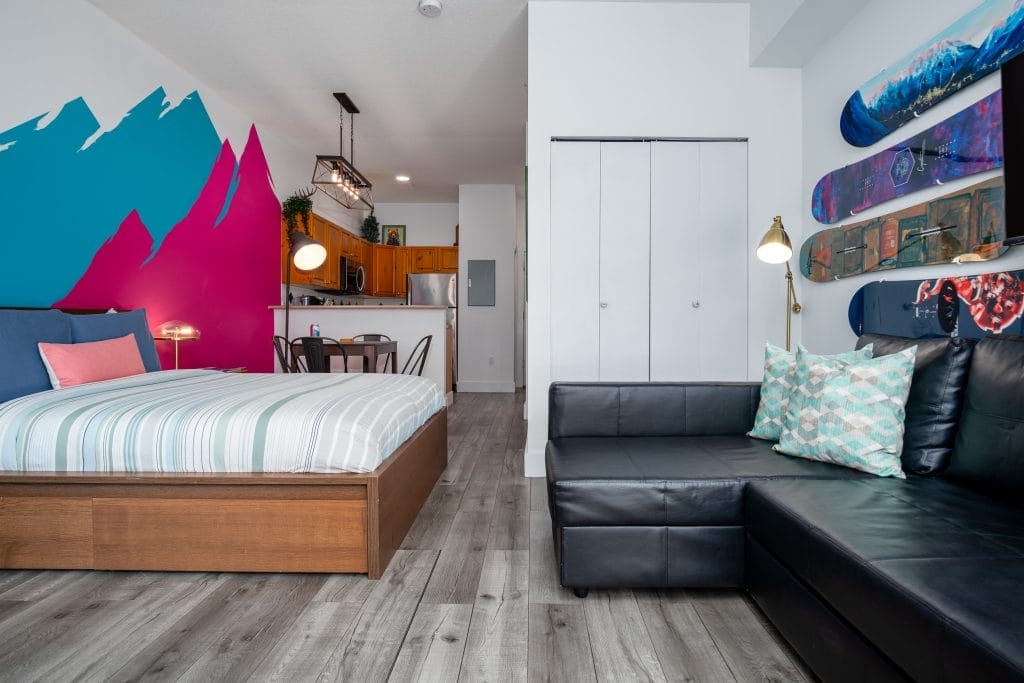 A modern studio apartment features a bed, black sectional sofa, wall art, and a kitchen area with dining table; the bright blue and pink accent wall with mountain design showcases our unique about us style.