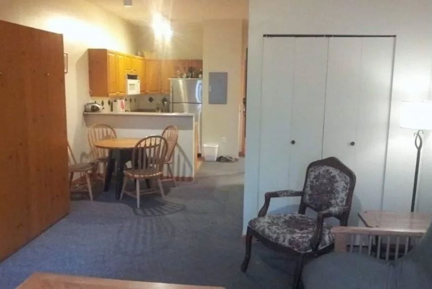 Open-plan living area with a small dining table and chairs, kitchen with wooden cabinets and stainless steel appliances, armchair, sofa, floor lamp—discover more about us through this stylish and inviting space.