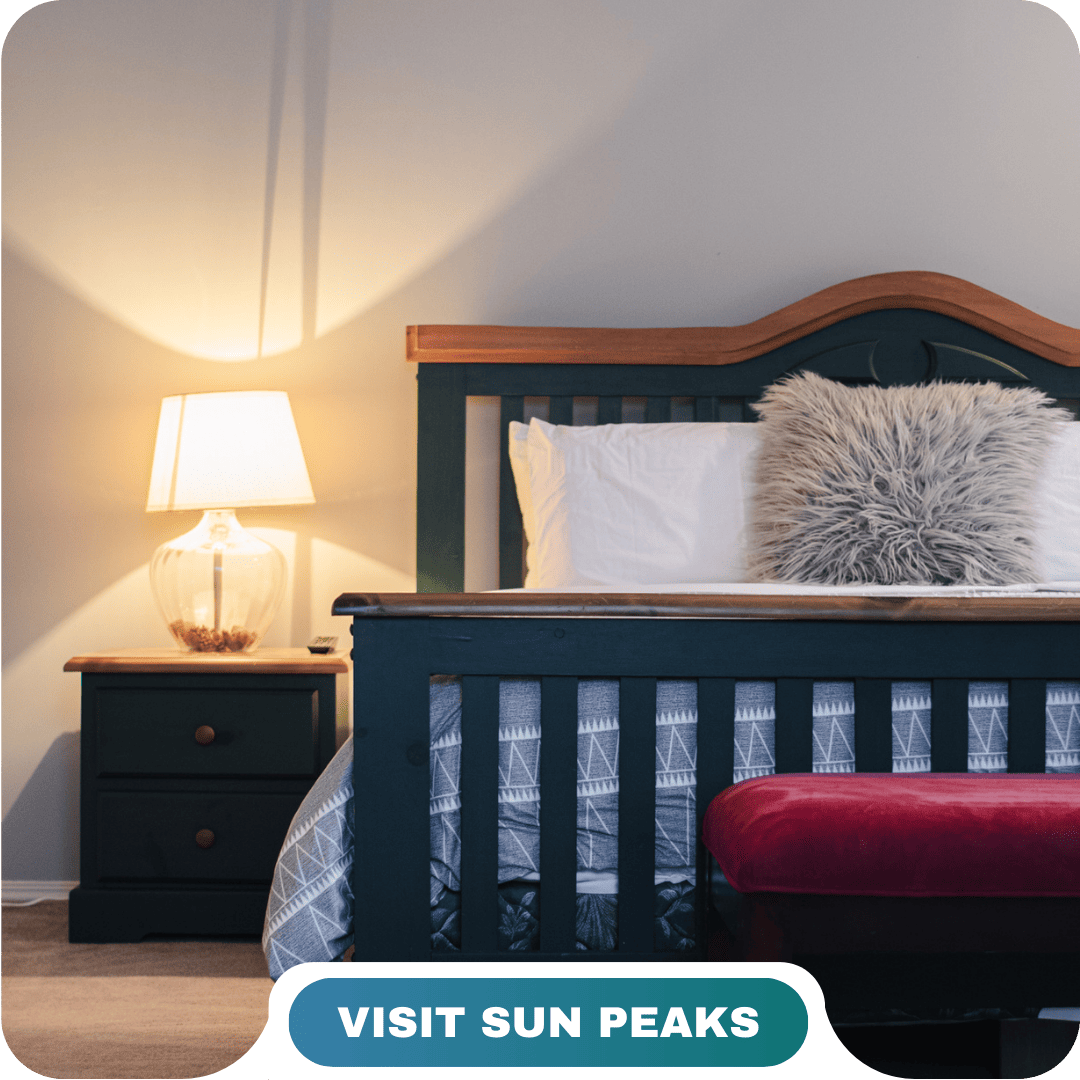 A neatly made bed with a gray fur pillow, wooden headboard, bedside table, and lamp. A red bench sits at the foot of the bed. Text reads "Visit Sun Peaks." Learn more about us and discover your perfect getaway.