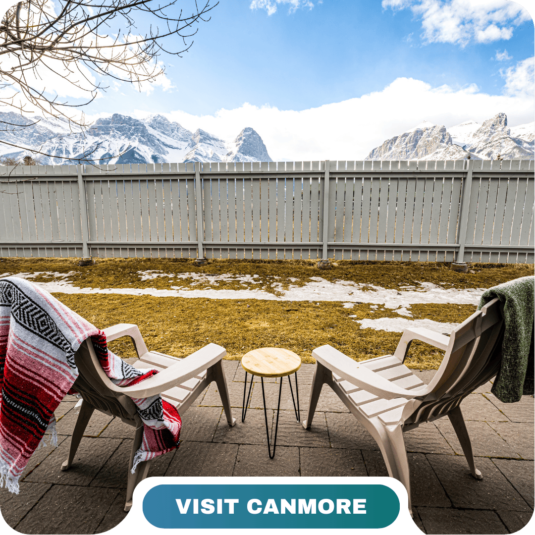 Two Adirondack chairs with blankets face a small table on a patio, overlooking a fenced yard with snow patches and snow-capped mountains in the background. Experience Canmore with Lifty Life.