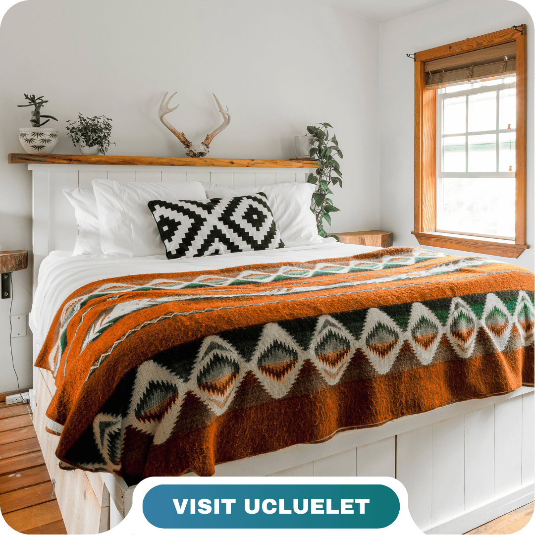 A neatly made bed with white linens, an orange patterned blanket, and a black-and-white cushion in a bright, rustic room; text reads "VISIT UCLUELET with Lifty Life.