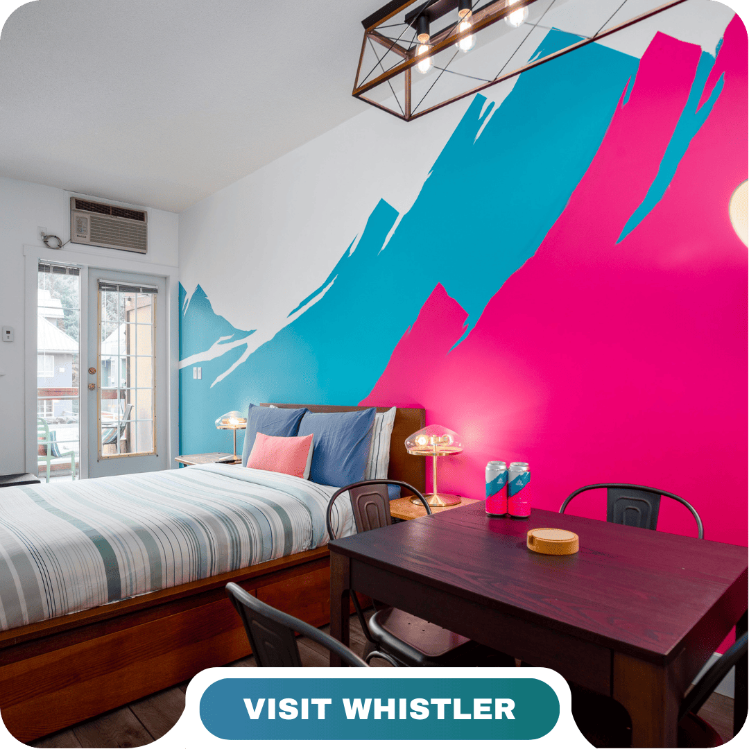 A modern bedroom with a striped bed, wall art of blue and pink mountains, a small dining table, and a "Visit Whistler" label at the bottom—perfect for enjoying Lifty Life comfort.