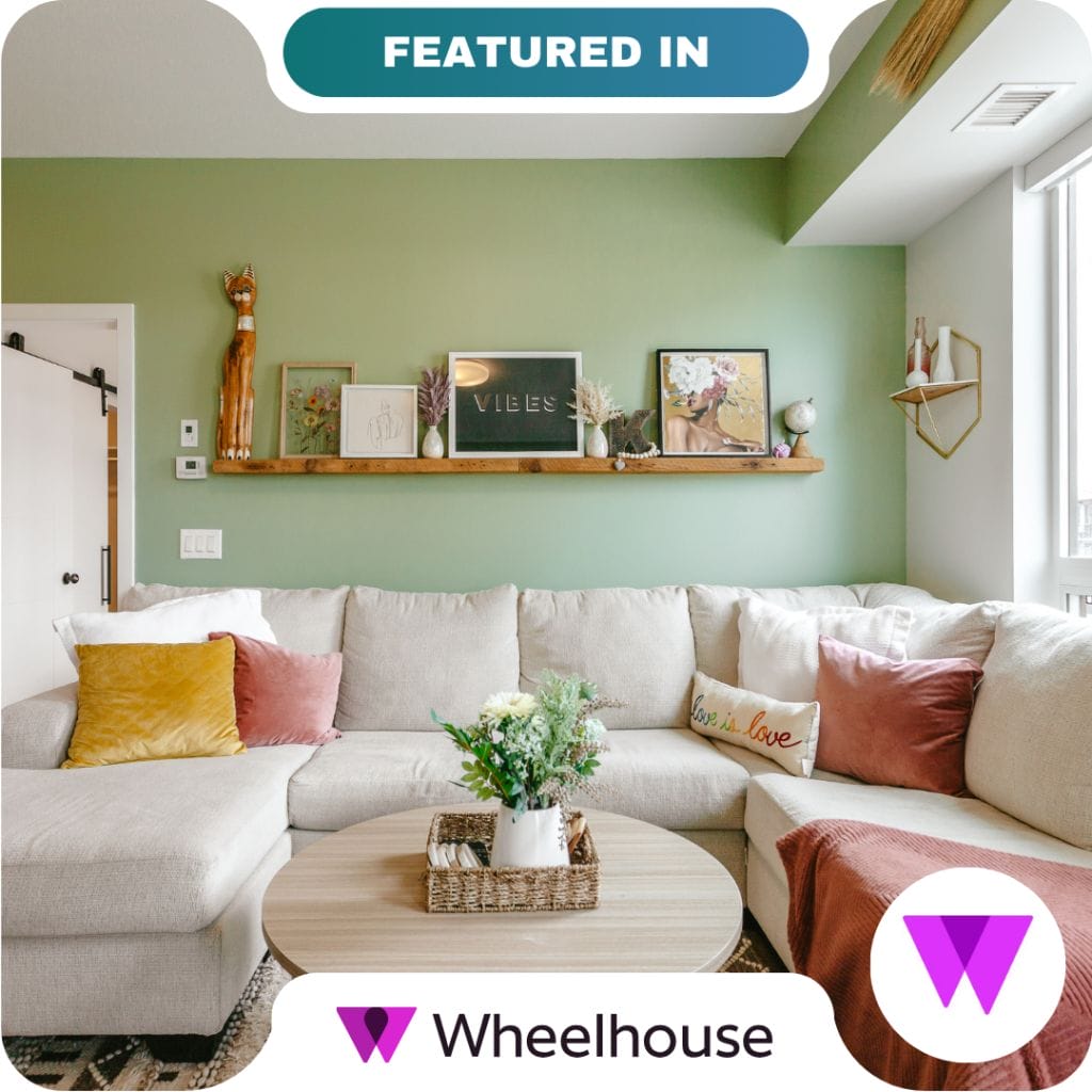 A modern living room with a light sectional sofa, colorful pillows, and a round coffee table with flowers. The green accent wall partners perfectly with art and decor arranged on a wooden shelf.