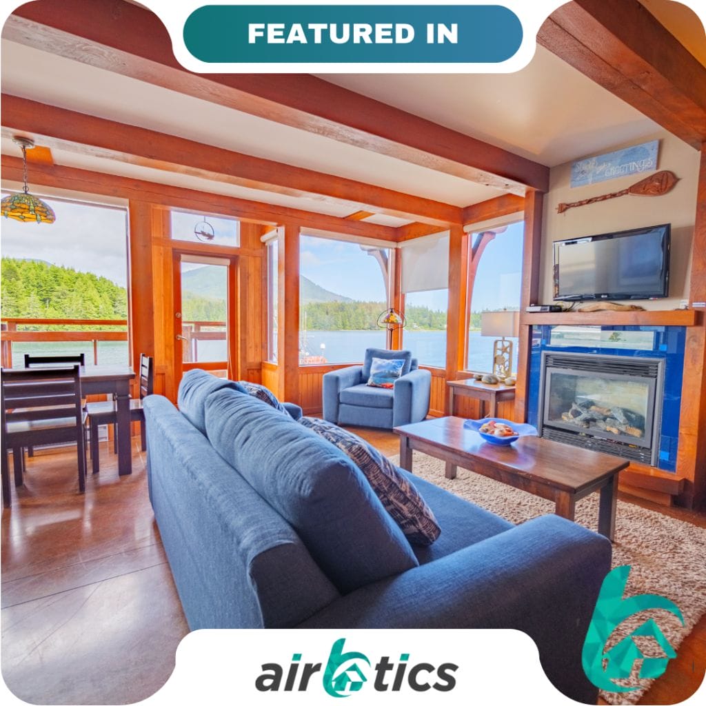 Modern living room with blue furniture, wooden accents, large windows overlooking trees and mountains, dining area, fireplace, and mounted TV. "Featured in Airbtics" at the top—perfect for partners seeking a cozy retreat.