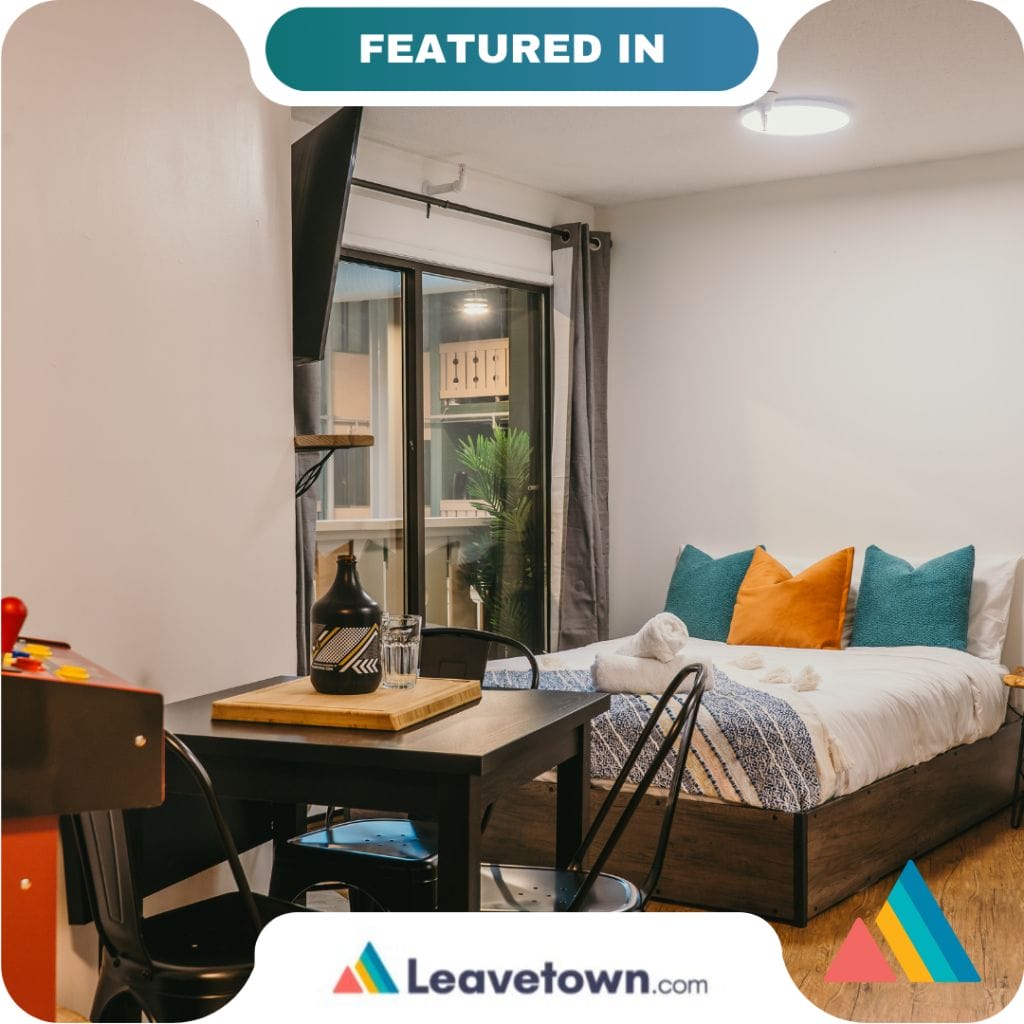 Modern studio apartment with a bed, small dining table, wall-mounted TV, and sliding glass doors leading to a balcony. Perfect for solo travelers or partners. Featured on Leavetown.com.