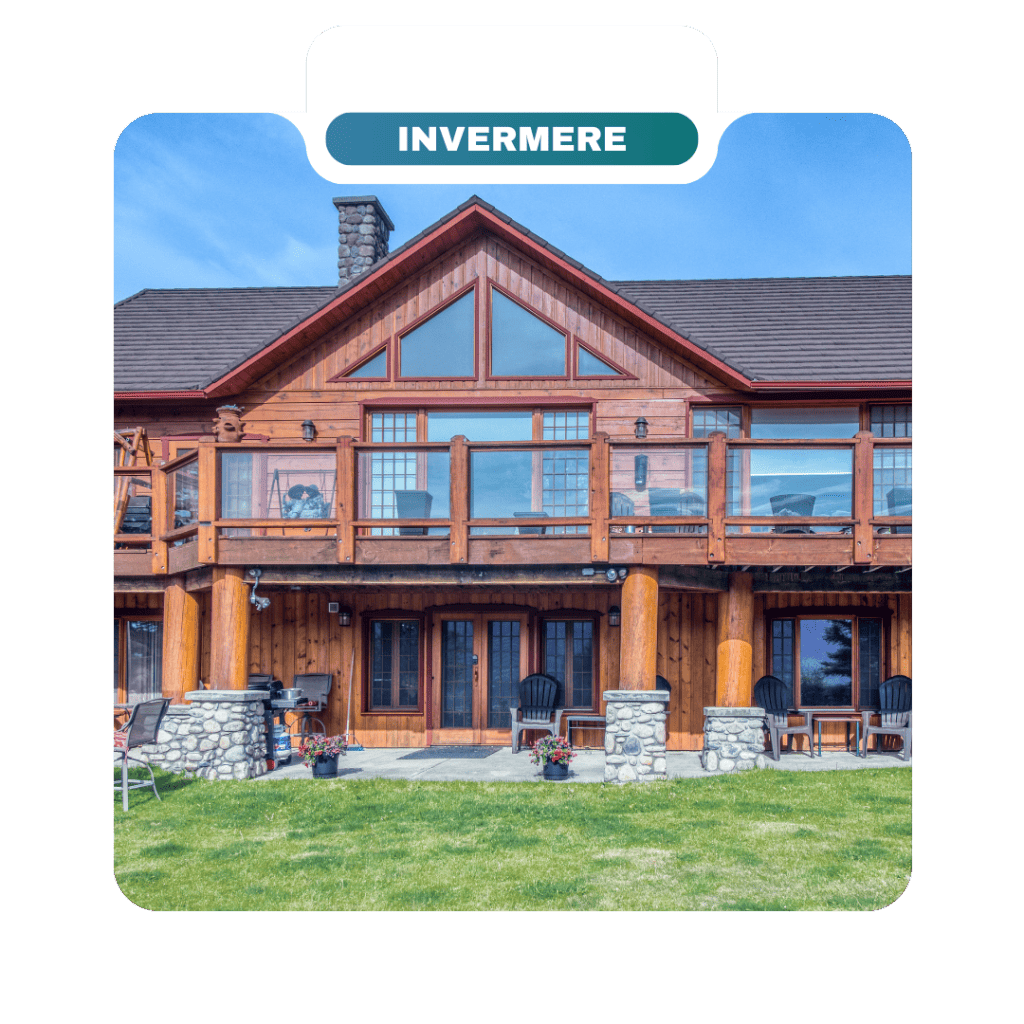 A two-story wooden house with large windows, stone pillars, a balcony with glass railing, patio chairs, and potted flowers on a grassy lawn is perfect for homeowners seeking beautiful destinations to unwind.