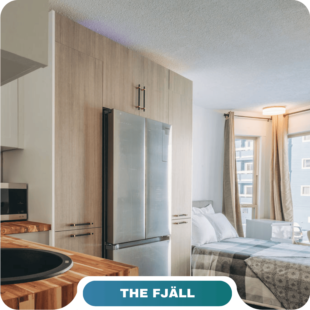 Modern studio apartment with a stainless steel refrigerator, wooden cabinetry, patterned bedding, and large windows with beige curtains—perfect for those seeking vacation rentals in Big White.