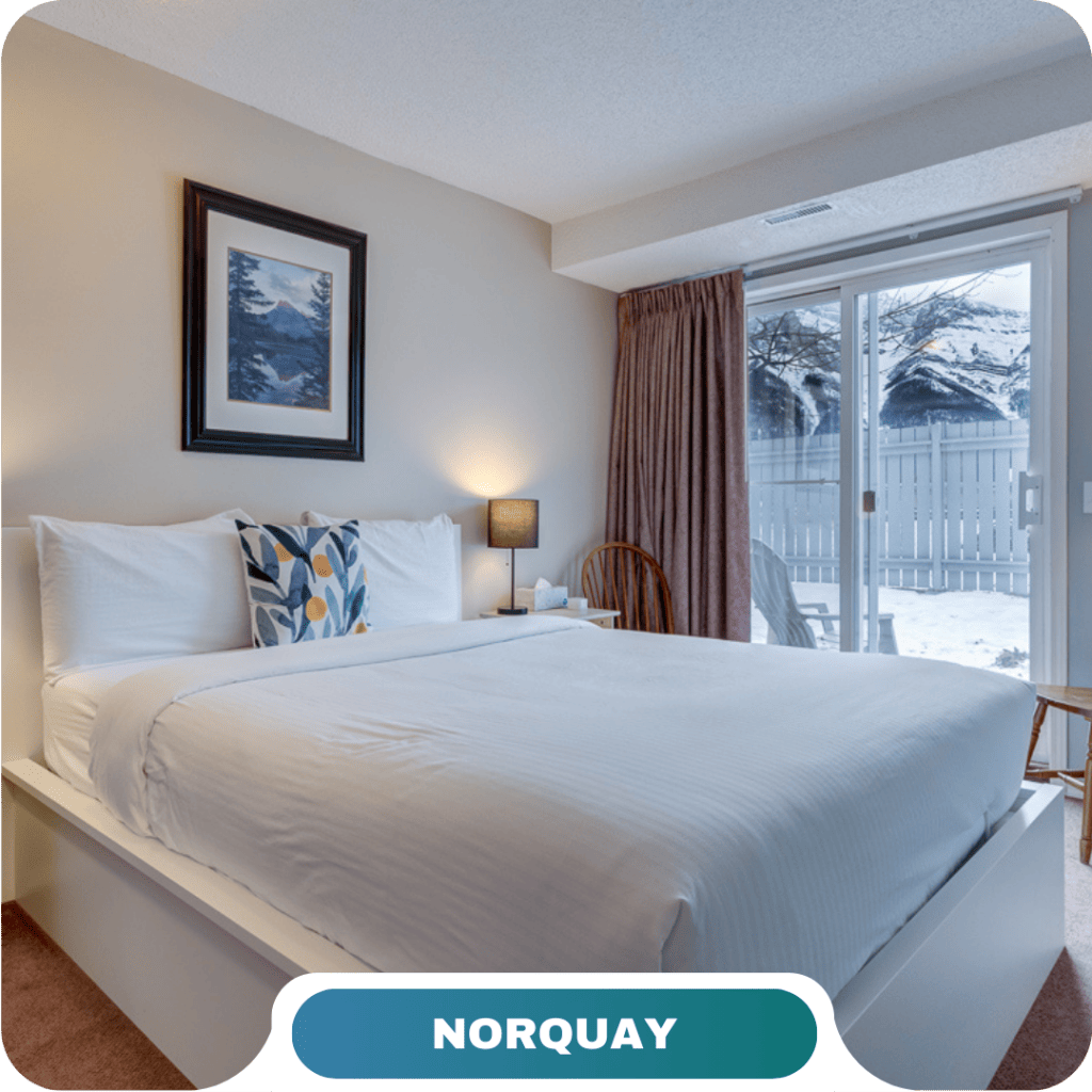 A neatly made bed in a bright room with a framed picture above, a lamp on the nightstand, and a sliding glass door opening to a snowy outdoor scene—perfect for vacation rentals in Canmore.