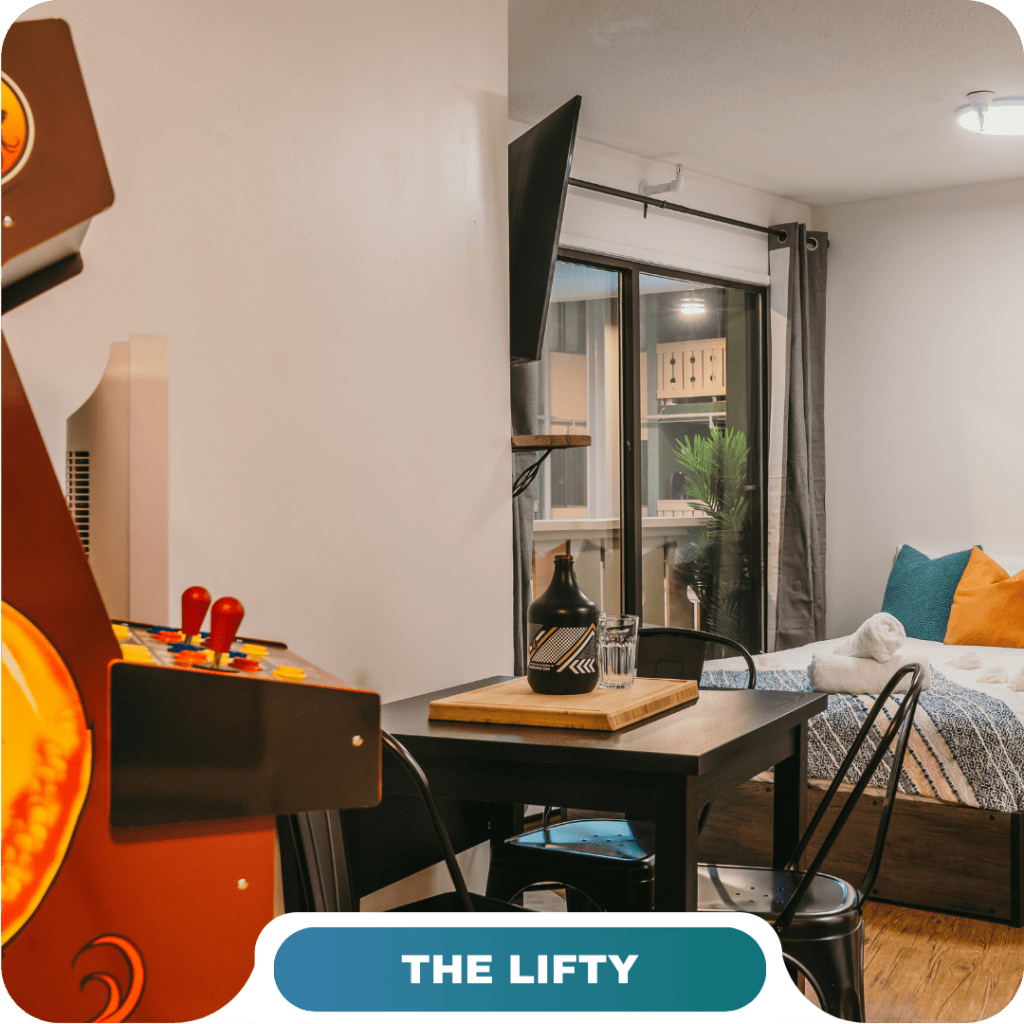 A modern studio featuring a bed, dining table, wall-mounted TV, large window, and an arcade game machine—the perfect option for vacation rentals in Big White.