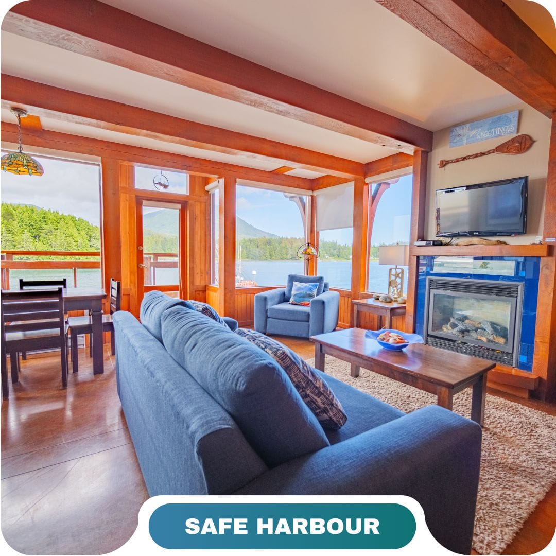 Bright living room with blue sofas, fireplace, dining area, large windows, and views of trees and mountains—perfect for vacation rentals in Big White; labeled "Safe Harbour" at the bottom.