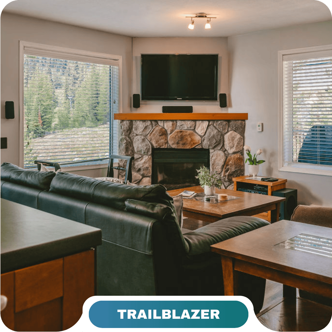 A cozy living room with a stone fireplace, wall-mounted TV, black leather sofa, and large windows showing a forested mountain view—perfect for vacation rentals in Big White.