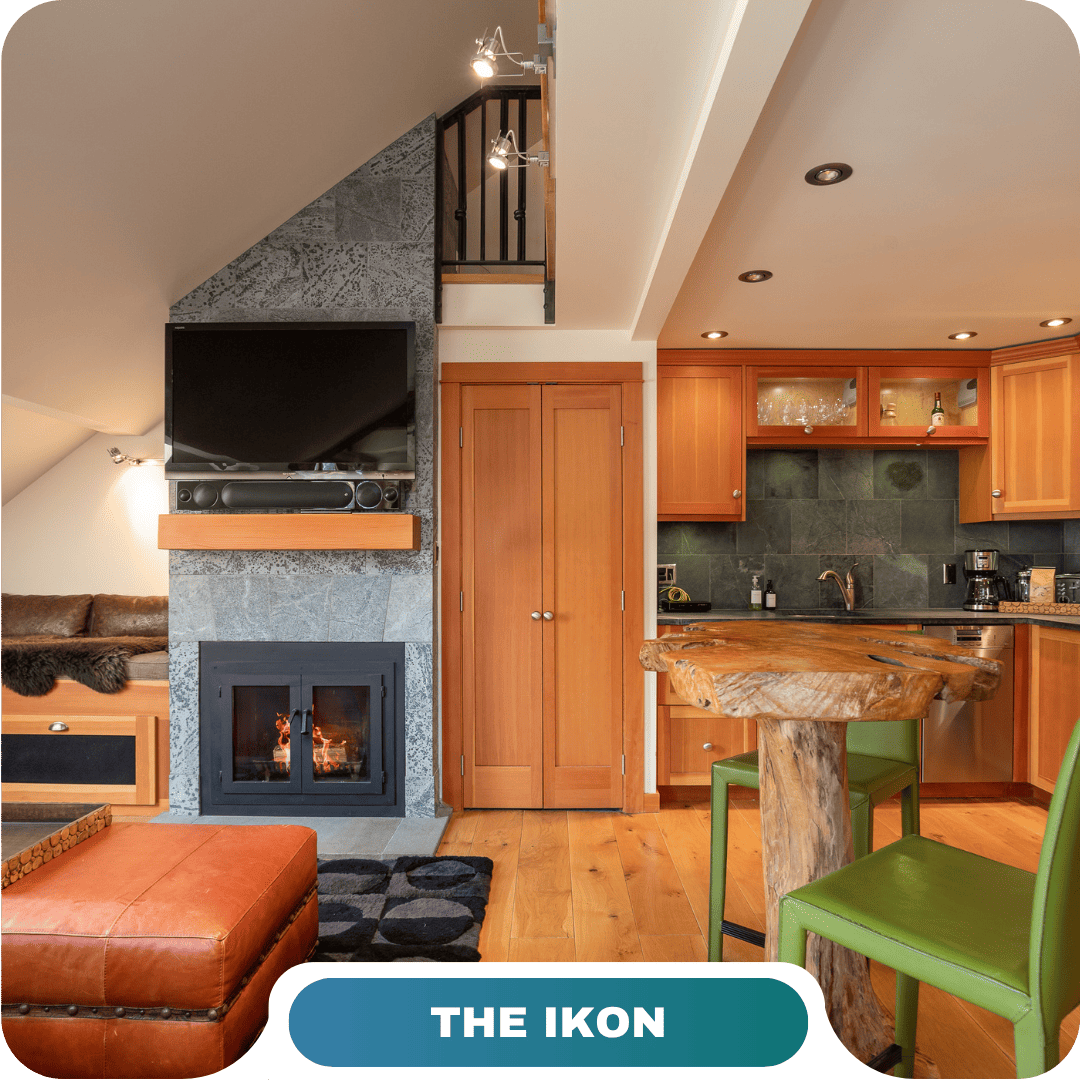 A cozy living area with a stone fireplace, wall-mounted TV, wooden furniture, a small kitchen, and a staircase—THE IKON offers an inviting retreat among the top vacation rentals in Whistler.