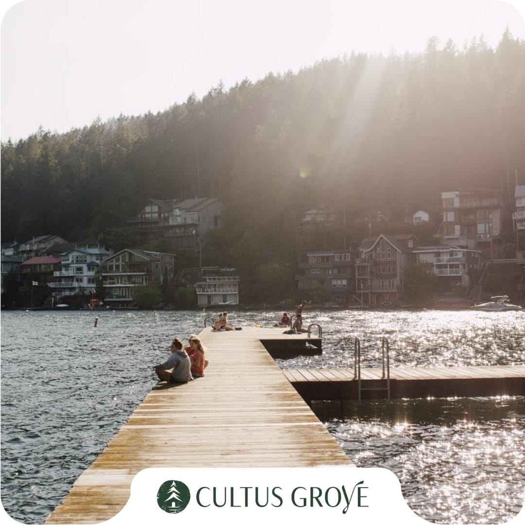 People sit and relax on a wooden dock extending into a lake, with houses and forested hills in the background under sunlight—ideal for those seeking vacation rentals in Cultus Lake. "Cultus Grove" logo is at the image’s bottom.
