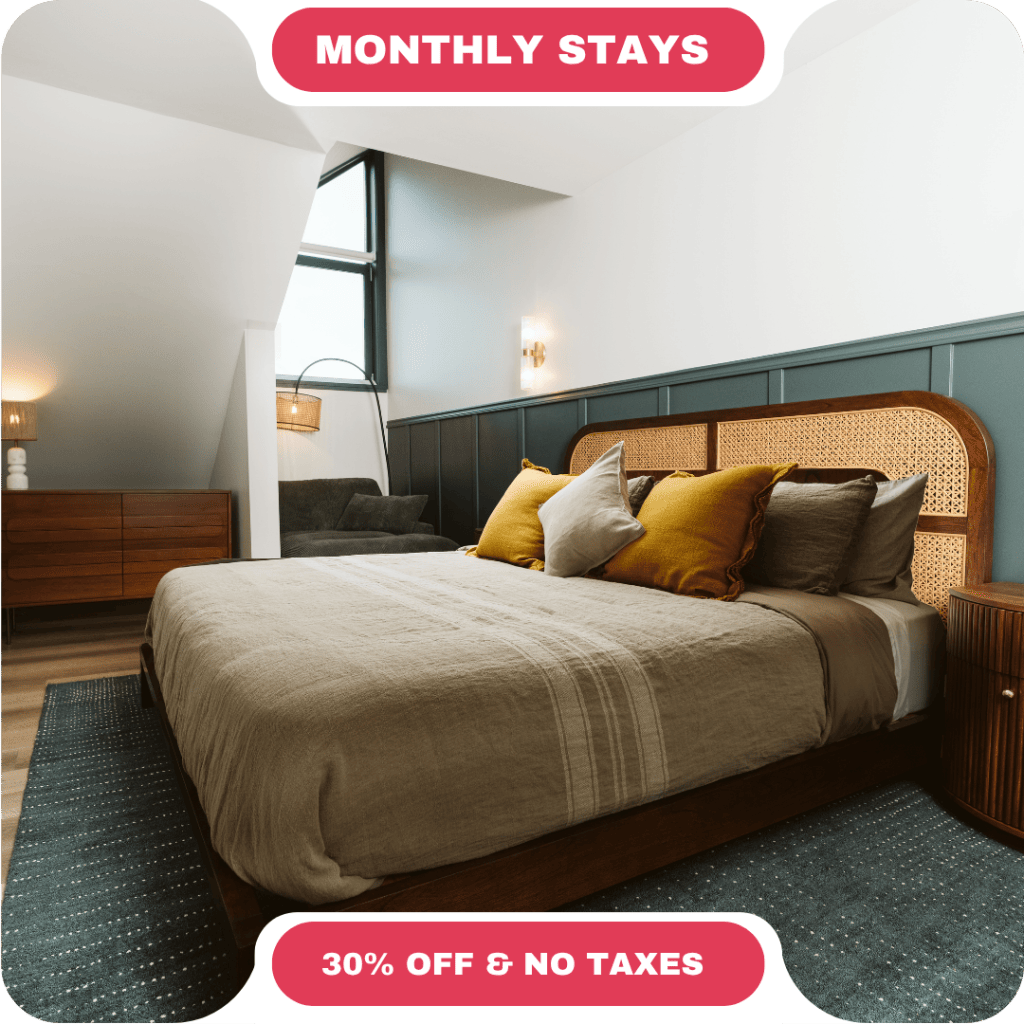 Modern bedroom with a large bed, decorative pillows, armchair, wooden furniture, and a large window—perfect for vacation rentals in Whistler. Text reads "Monthly Stays" and "30% Off & No Taxes.