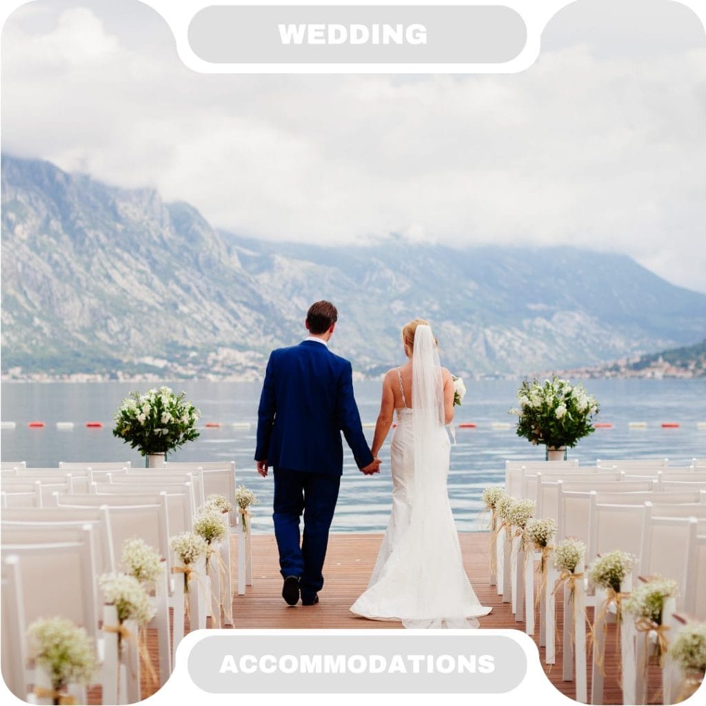 A bride and groom walk hand in hand down an outdoor aisle toward a lake, with mountains in the background and white chairs decorated with flowers—an atmosphere as inviting as the finest furnished rentals.
