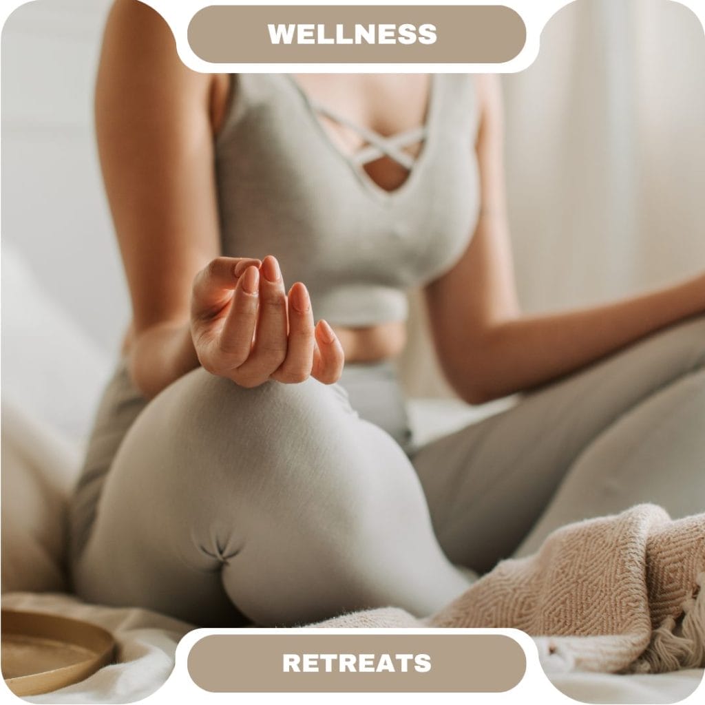 A person in grey activewear sits cross-legged on a bed in a stylish furnished rental, meditating with one hand resting on their knee. The image is labeled “Wellness Retreats.”.